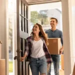 Happy first-time home buyers entering their new house with moving boxes using home buyer grants assistance